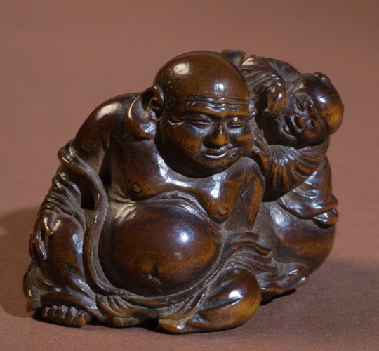 SHUGETSU: LUCKY GOD HOTEI AND BOY: Netsuke, wood. Japan, 19th cent. The large bellied lucky god is sitting and leaning against a sack. He is laughing only slightly and amusingly showing a little pain in his face, as the boy sitting on 