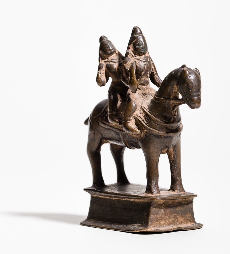 DEITY WITH SPOUSE ON HORSEBACK: Bronze. India, approx. 16th cent. A standing horse on a tiered base, upon which the deities are seated, in front the male holding sword and reins, behind him the little female. Presumably the deity Kh