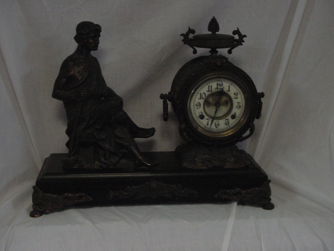 New Haven Open Escapement Clock: A barefoot seated draped figure is seated and holding a sledge hammer handle in his right hand, looking at an open escapement beveled glass door clock with an urn decoration on top of the case, black 
