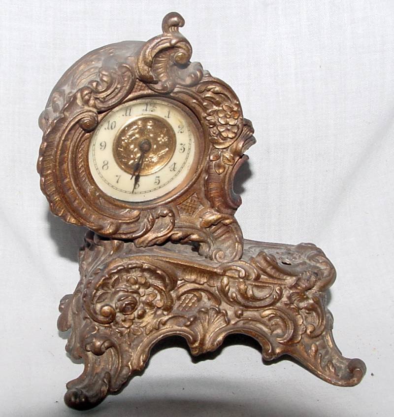 Ornate Brass Desk/Shelf Clock: Probably 1880-1910 era, unsigned, recessed center dial with enameled face, extremely ornate case with something missing from the side of the top of the base, scroll feet, 4 1/2" long and tall. The clo
