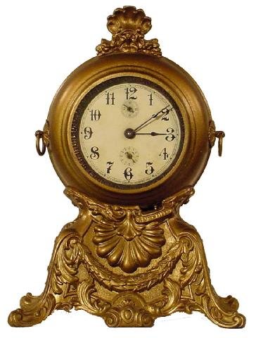 Gilbert Novelty Mantel Clock: Cast Iron Case w/Garland On Base & Scroll Work, Stylized Shell Below Main Clock, The Windup Clock Resembles A Large Pocket Watch On A Stand, Signed On Center Of Face W. L. Gilbert Clock Co., Winsted,