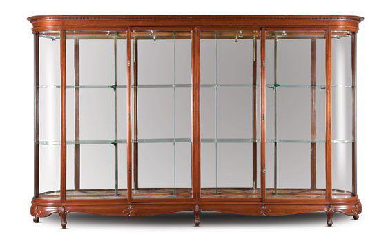 One of a Pair of Large Mahogany Display Cabinets: OFFERED WITHOUT RESERVE English, circa 1900. With an overhanging molded cornice, above four glazed doors, opening to glass shelves, with concave glazed sides, the shaped skirt carved with leaf sprays,