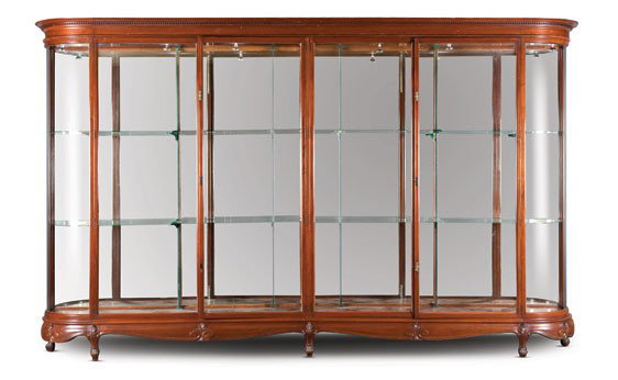 One of a Pair of Large Mahogany Display Cabinets: OFFERED WITHOUT RESERVE English, circa 1900. With an overhanging molded cornice, above four glazed doors, opening to glass shelves, with concave glazed sides, the shaped skirt carved with leaf sprays,