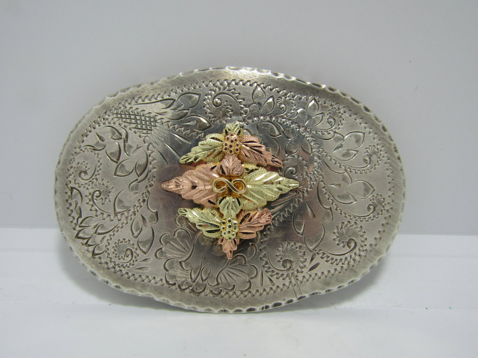 LANDSTROM'S 10K GOLD & STERLING BLACK HILLS BUCKLE: ORNATE VINTAGE LANDSTROM'S BLACK HILLS GOLD BELT BUCKLE. CHASED SCROLLWORK AND LEAF DESIGN. THICK 10K GOLD LEAF ON FRONT. SIGNED L. TESTED & MARKED STERLING SILVER & 10K GOLD. MEASURES 3" X 2". FITS 1