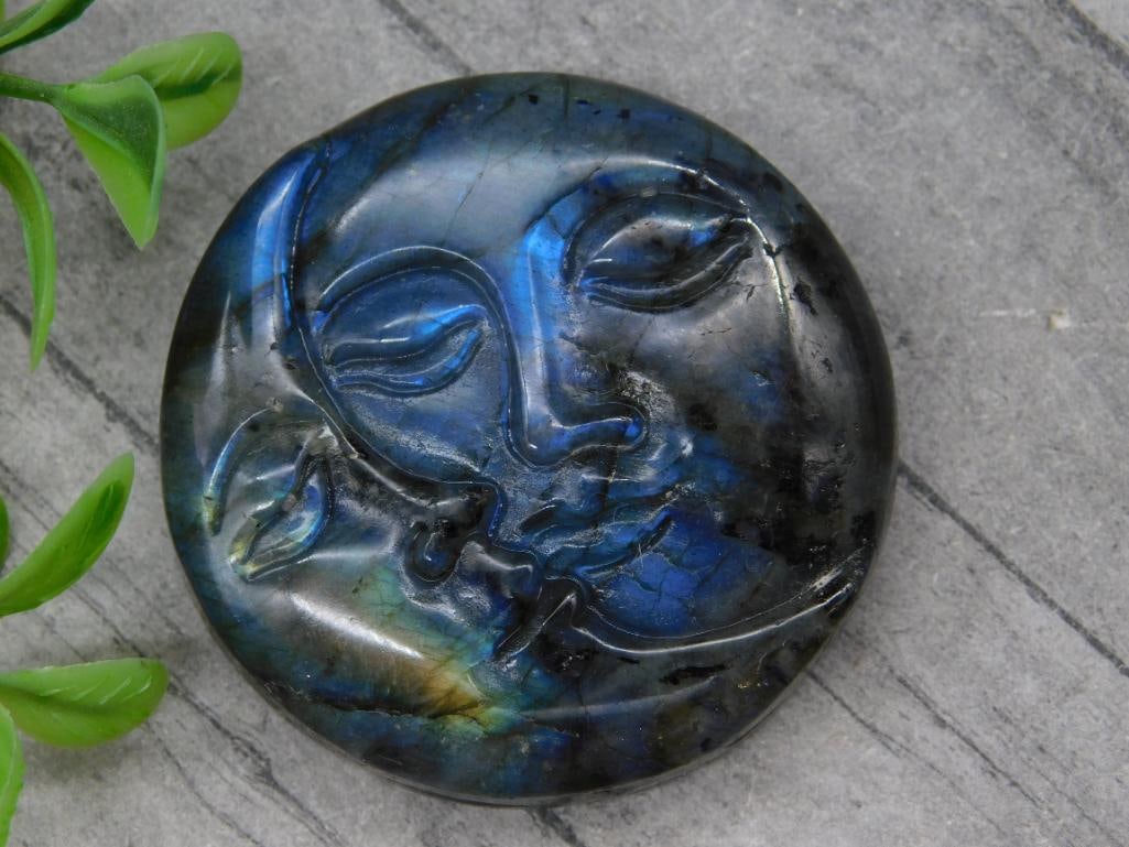 Carved Labradorite Gemstone With Face Design: Round labradorite gemstone featuring a carved design of a serene human face. Displays vibrant iridescent hues characteristic of labradorite. Weighs approximately 43 grams. The size is shown in compari