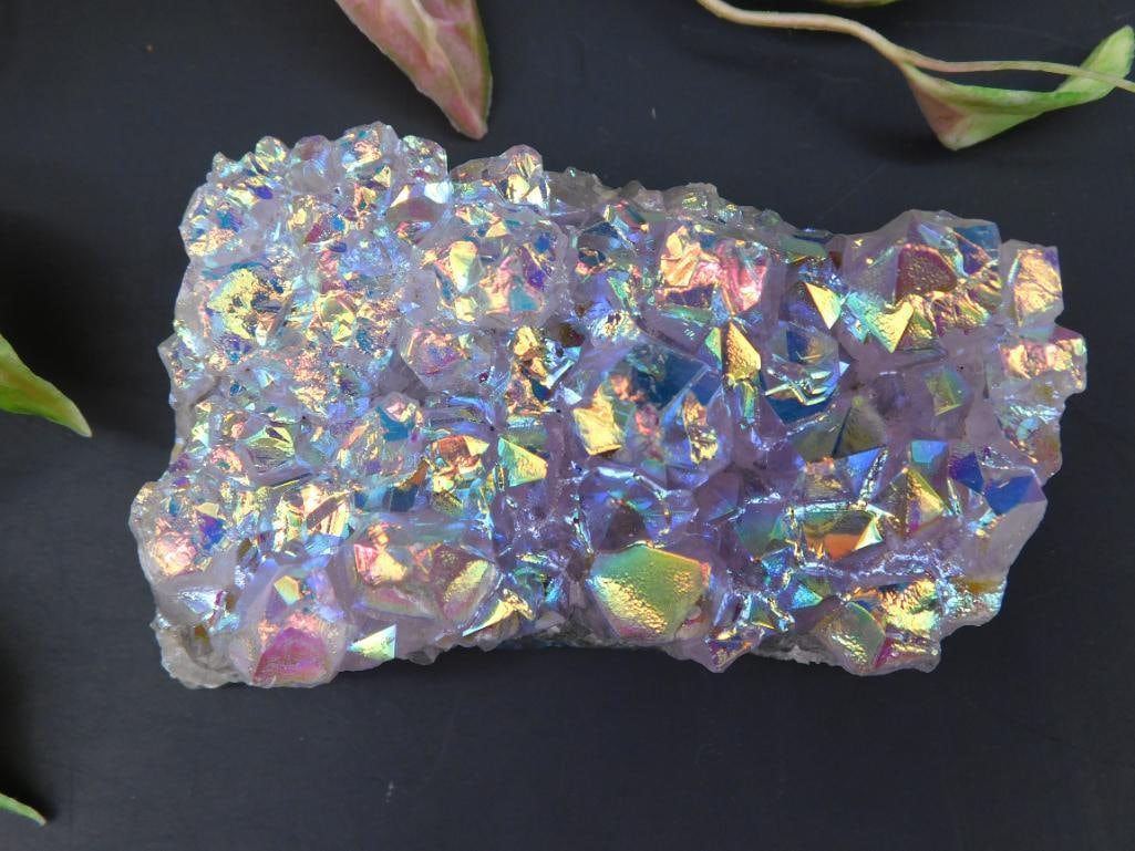 Angel Aura Amethyst: This is a sparkling, iridescent quartz cluster with a reflective coated finish, creating a multicolored display. The stone comprises angular crystal formations, providing a three-dimensional texture.