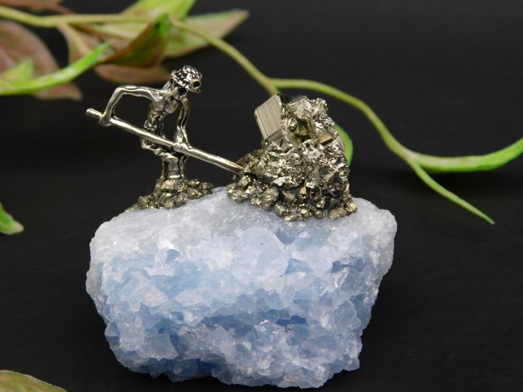 Miner Figurine on Blue Crystal Rock Display: This item features a miniature miner figurine sculpted in metal, posed working with a pile of rock. The figurine is mounted atop a natural blue crystal rock base, providing a unique contrast between t