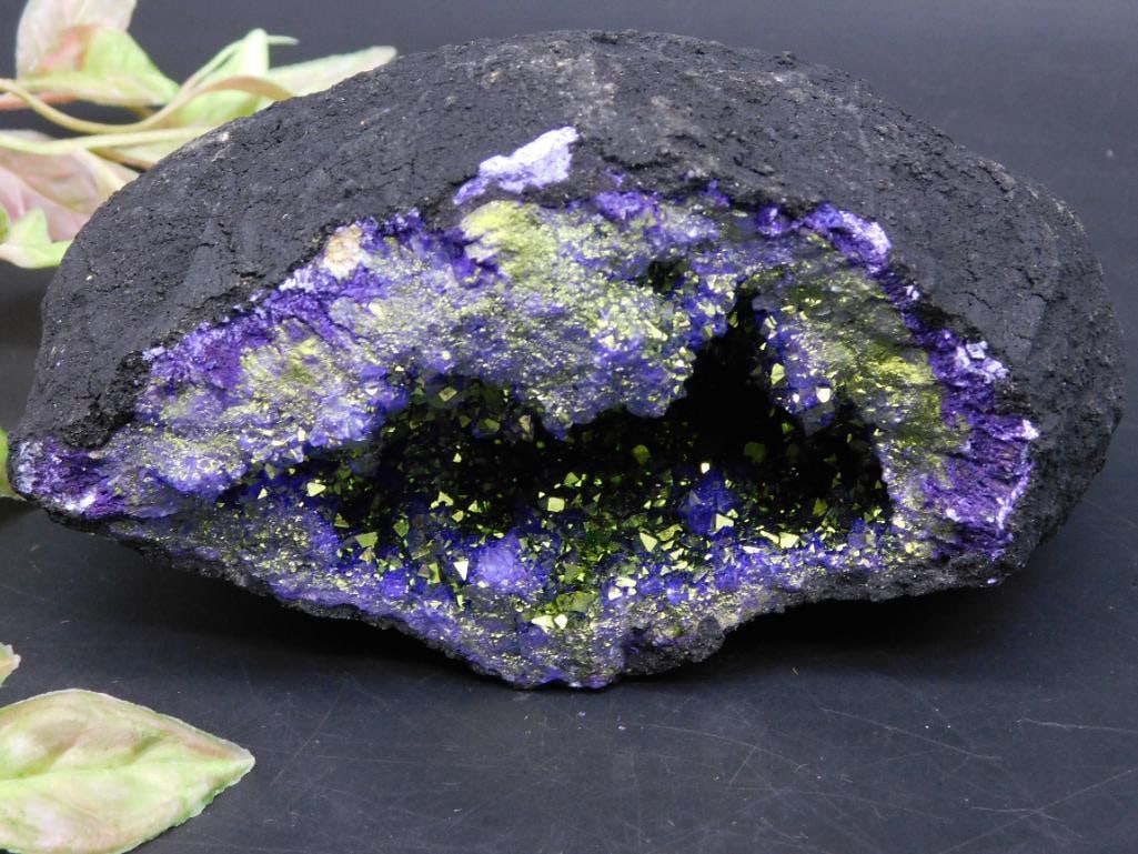 Purple and Green Mineral Geode Display Rock: Purple and green mineral geode with vibrant interior. Features dark exterior surrounding an open cavity adorned with sparkling mineral formations. Weighs 1 pound 7 grams as indicated on the tag. The m