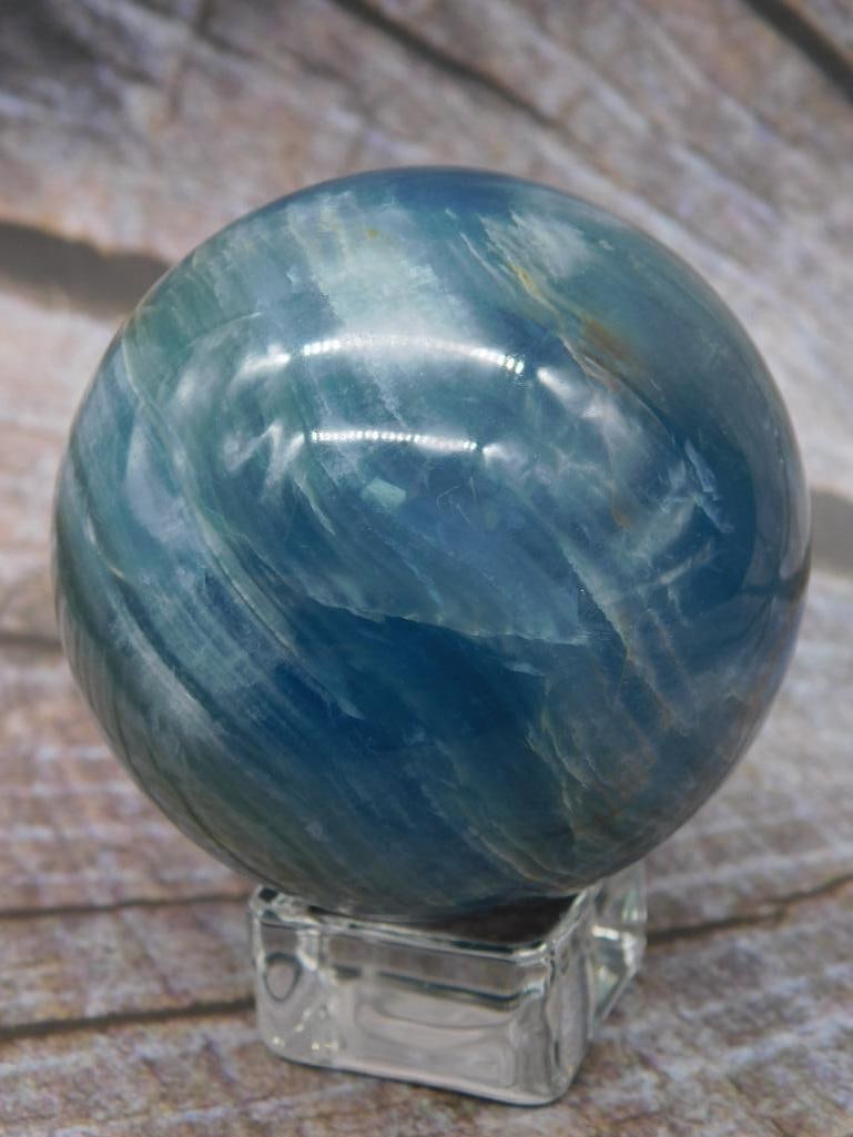 Blue Mineral Sphere: A polished blue mineral sphere with striking patterns, displayed on a transparent rectangular stand. The sphere has a unique swirl design throughout, ideal for decorative or collection purposes. Inclu