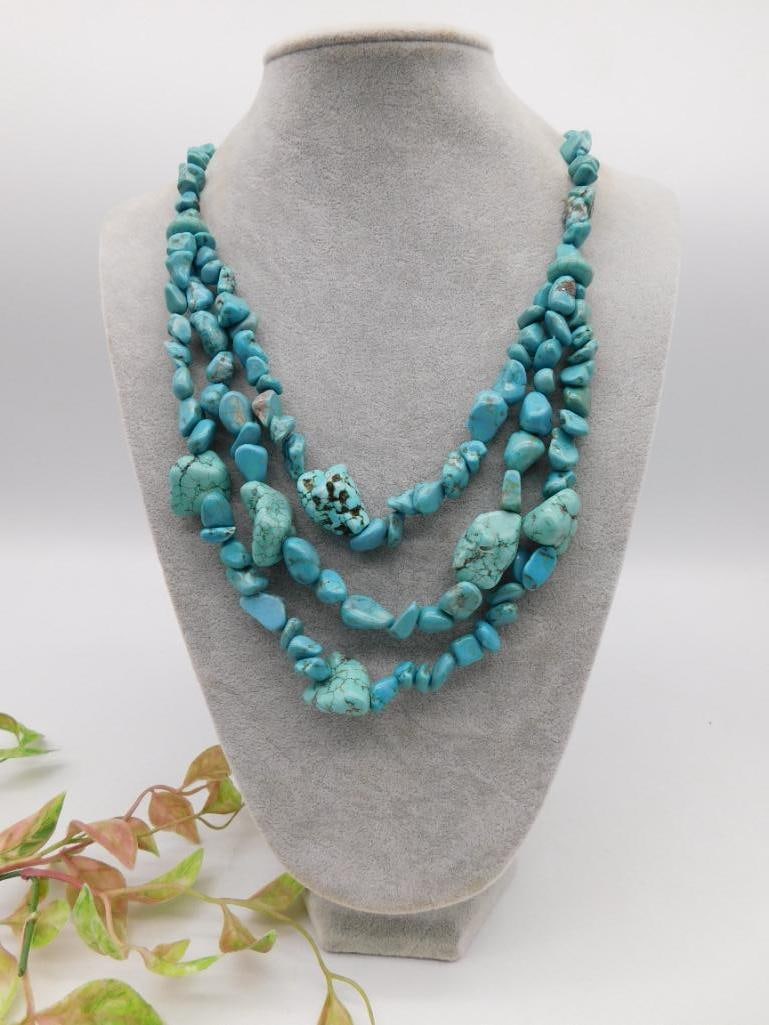 Three-Strand Turquoise Bead Necklace: A three-strand turquoise bead necklace featuring irregularly shaped turquoise stones with varying shades and veining patterns. The strands are interconnected and displayed on a gray jewelry bust. The 
