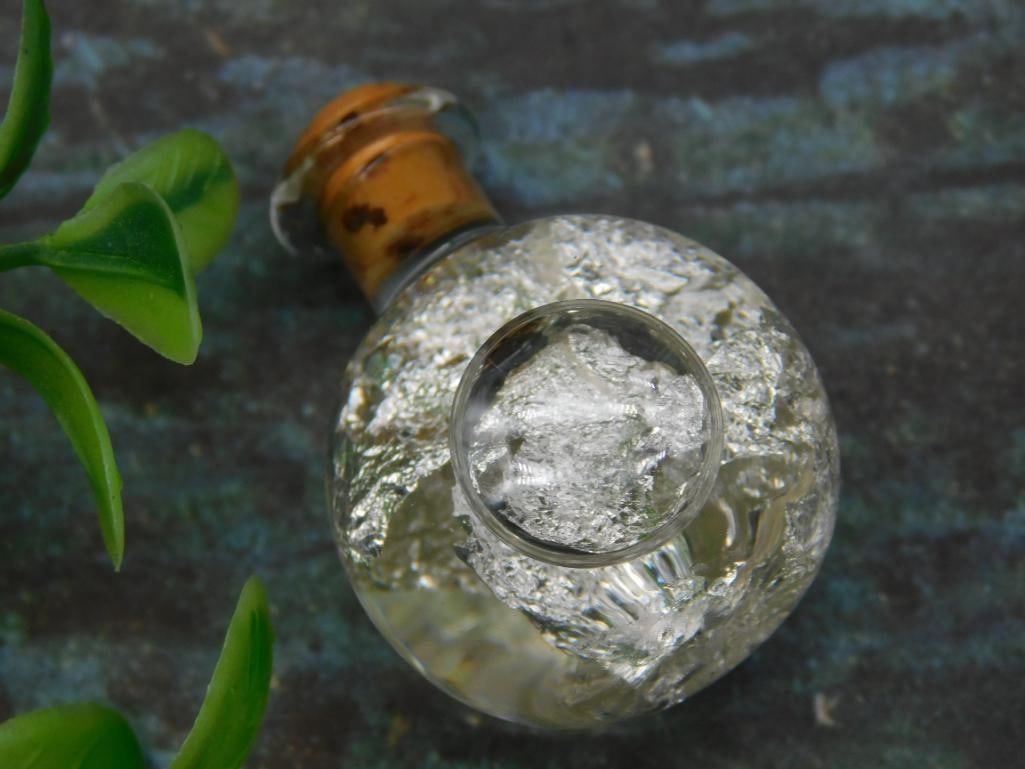Glass Bottle with SILVER FLAKES and Cork Stopper: Small round glass bottle containing a clear mineral specimen. Features a removable cork stopper and transparent body. Bottle measures approximately the size of a U.S. quarter for scale. Weighs 10 gram