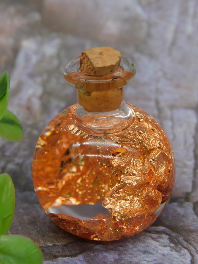 Small Glass Bottle with COPPER FLAKES: A small round glass bottle containing gold-toned leafing, sealed with a cork stopper. The bottle is compact and spherical in shape, as shown next to a quarter for scale. The gold-toned leafing creates