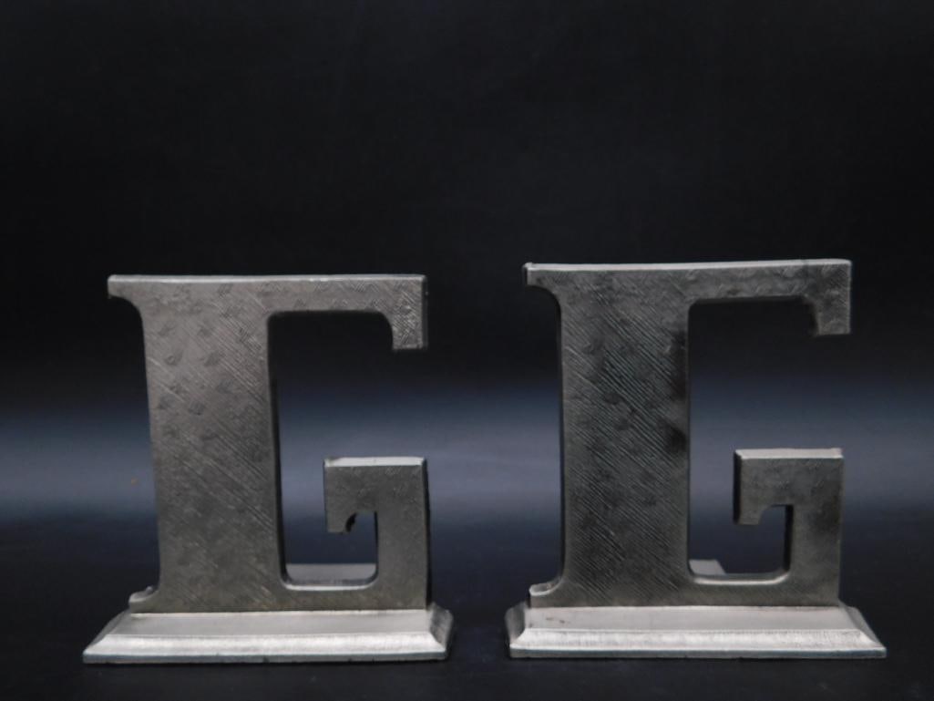 Pair of Metal Bookends in G Shape Design: This pair of metal bookends feature a bold 'G' shape design. Crafted to hold books in place, each piece is constructed of metal and has a textured surface finish. A manufacturer's mark ? VIP is presen