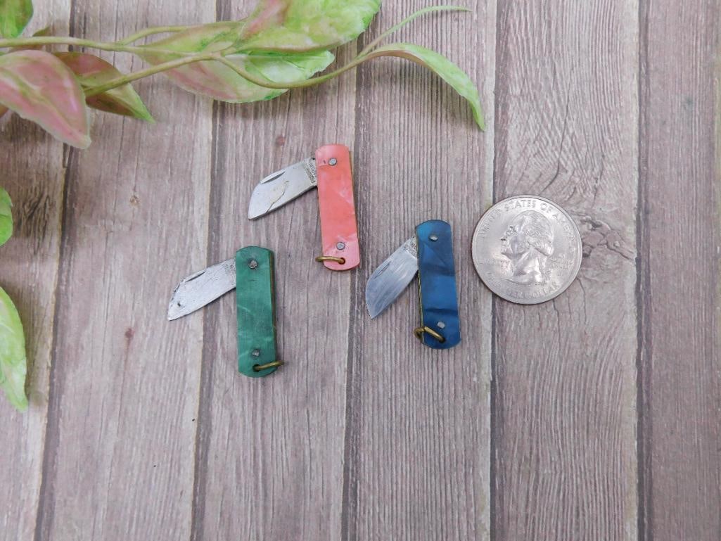 Set of 3 Miniature Folding Pocket Knives: This is a set of three miniature folding pocket knives in green, red, and blue finishes. Each knife features a small folding blade and a brass pivot point. Compact size, displayed next to a coin and r