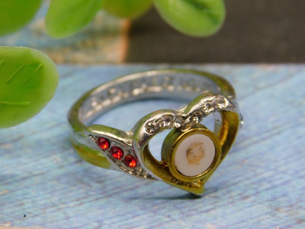 Heart-Shaped Ring With Red Stones and Circular Design: Women's ring featuring a heart-shaped design with small red stones embedded on the band, encasing a circular detail at the center. Silver-tone metal band with engraved detailing. Size 7, as indicated.