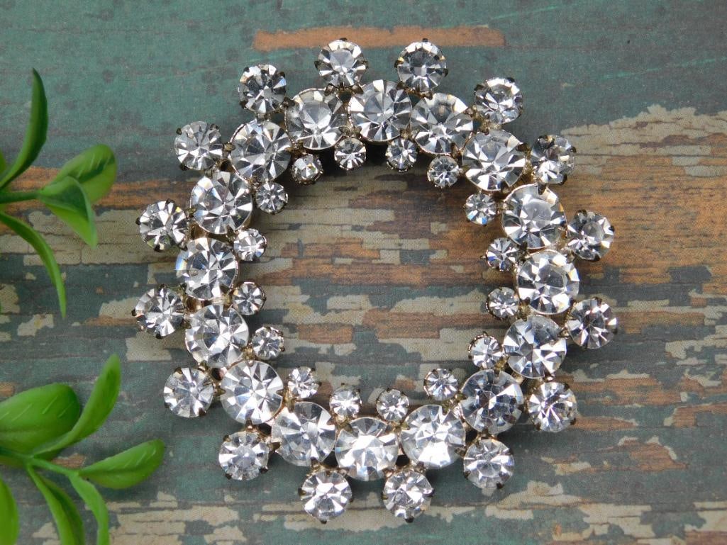 Circular Rhinestone Brooch with Silver-Tone Backing: This is a circular rhinestone brooch featuring clear, faceted rhinestones arranged in a layered design. The back has a silver-tone finish and a secure clasp mechanism for attachment. The brooch is sho