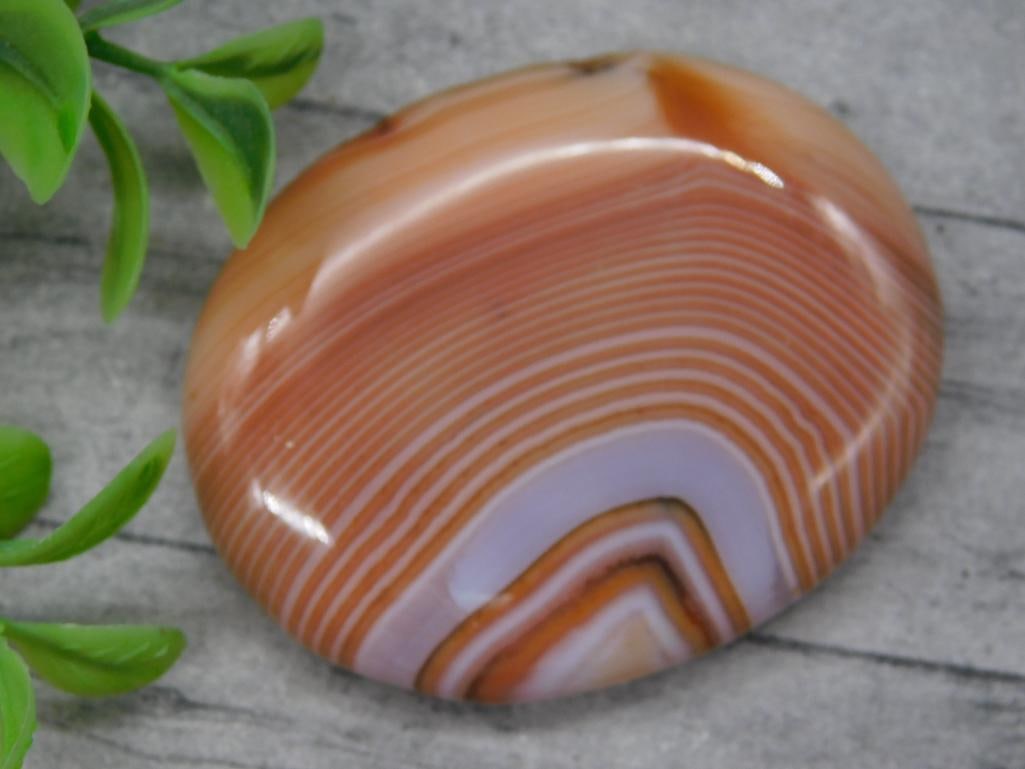 Polished Banded Agate Stone: A polished banded agate cabochon with vibrant striations of reddish-orange, white, and beige layers, creating a striking natural design. Oval-shaped with a smooth finish, suitable for display or craft