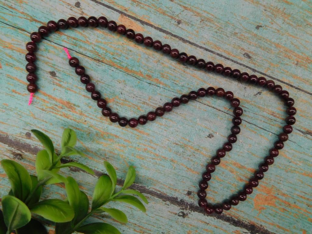 Strand of Garnet Beads Rock Stone Lapidary Specimen: - A single strand necklace composed of dark red beads. - Beads are spherical in shape with an even finish. - Necklace length is approximately visible against a standard ruler for scale. - Appears to b