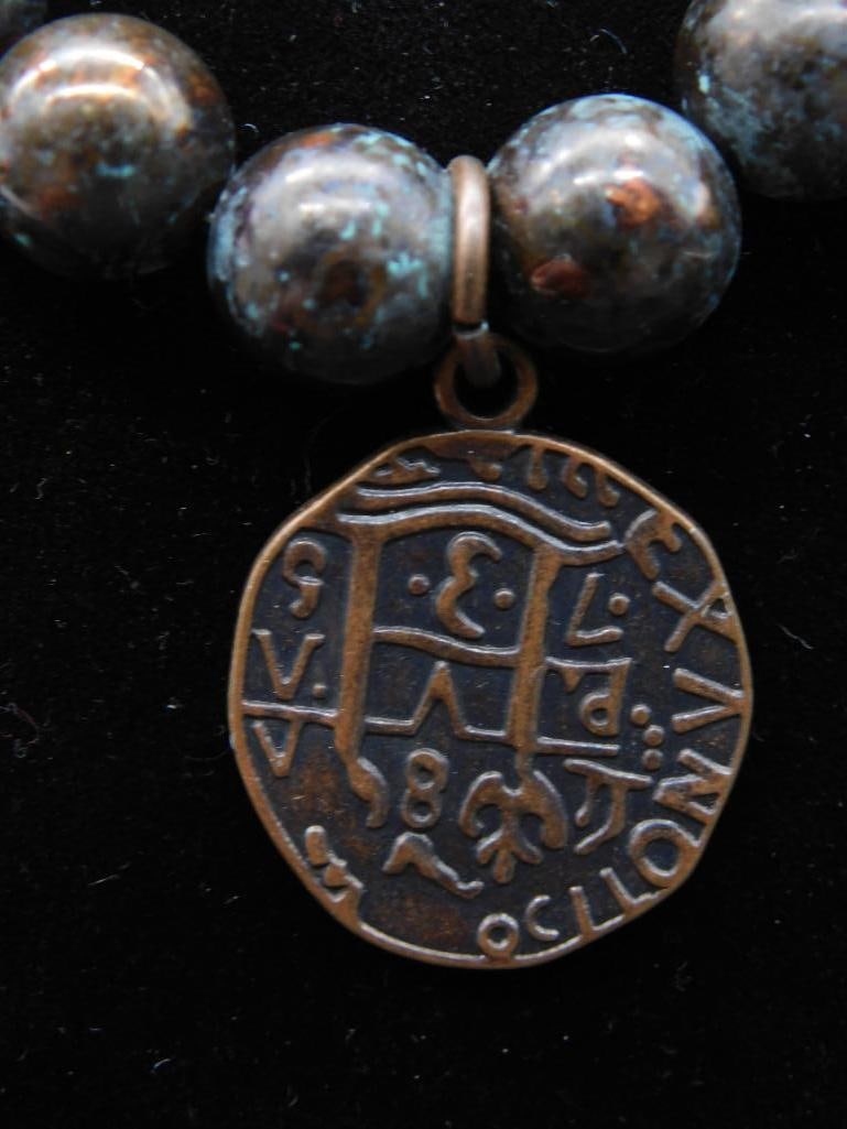 Necklace With Beads and Coin-Style Pendant: A necklace featuring spherical beads and a pendant reminiscent of a coin. The bead design exhibits tones suggestive of patina, offering a distinctive aged look. The clasp and bead threading appear int