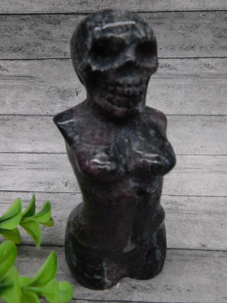 Carved Stone Figurine Featuring Torso and Skull Motif: Carved stone figure displaying a stylized torso with visible skull details. Made from polished stone with dark shading. Weighs approximately 261 grams. Includes auction lot information card.
