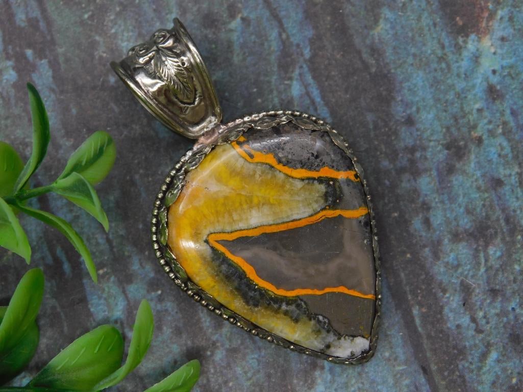 Bumblebee Jasper Teardrop Pendant with Intricate Tooling Rock Stone Lapidary Specimen: This teardrop-shaped pendant features a polished stone with bold orange and yellow accents set in a decorative metal frame. The back displays an intricate engraved design, and the hanging loop is styl
