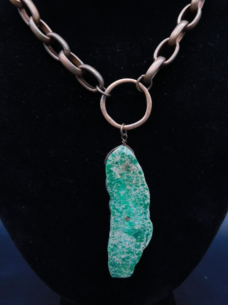 Copper Chain Necklace With Green Stone Pendant: Copper-tone chain necklace featuring a large green stone pendant. The chain has circular links connected to a round loop that attaches the irregularly shaped green pendant. Suitable as a decorative pi