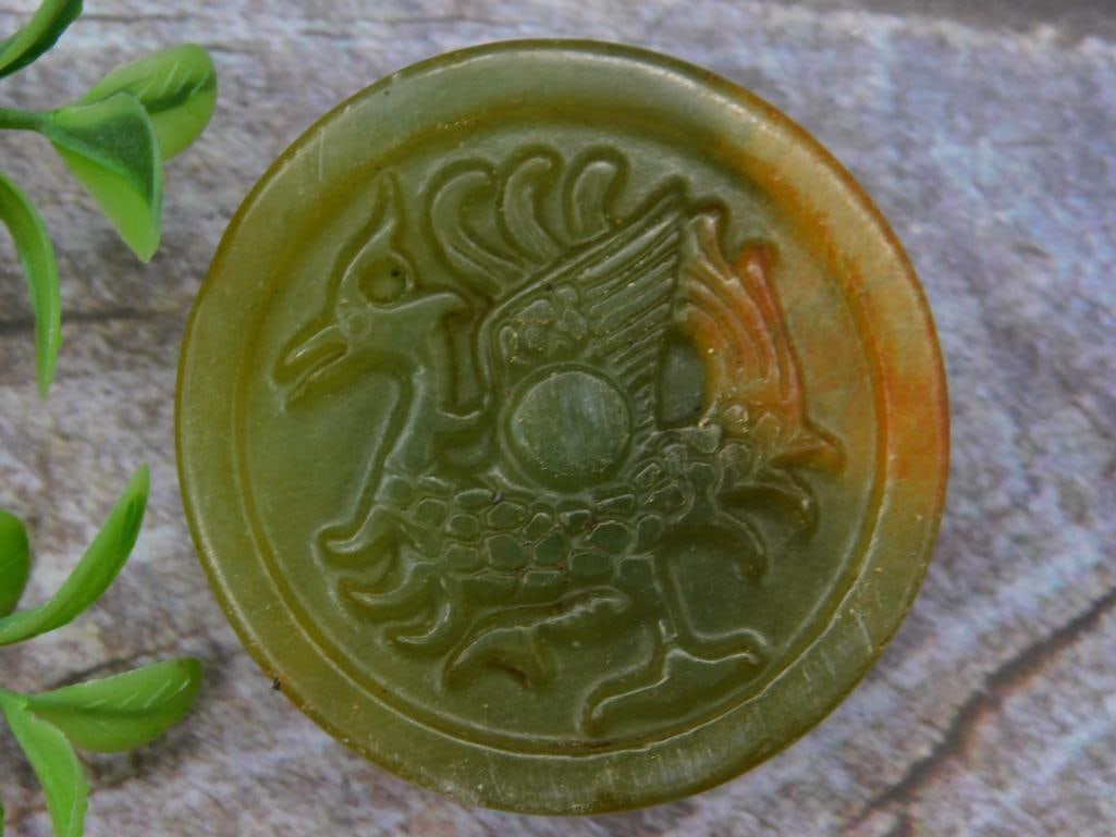 Carved Jade Stone Medallion with Animal Motifs: Carved green stone medallion featuring a winged creature on one side and a dragon-like design on the other. Appears to be circular in shape with detailed craftsmanship. Includes a comparative size ref