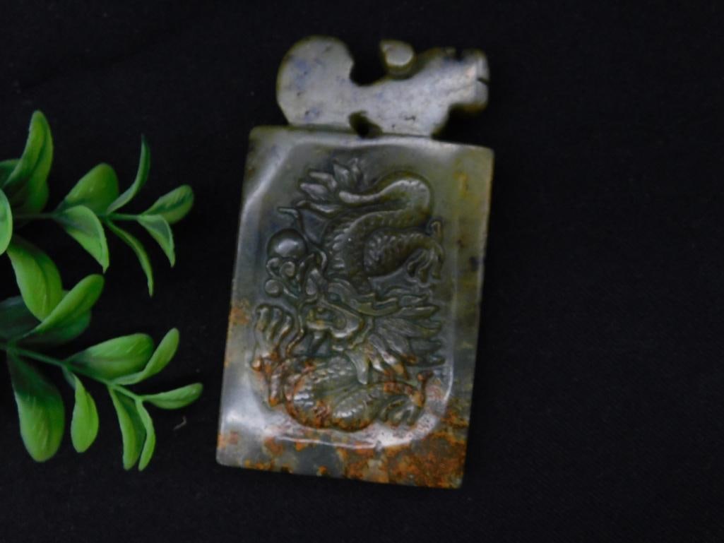 Carved Jade Pendant Featuring Dragon Motif Rock Stone Lapidary Specimen: Rectangular carved jade pendant with detailed dragon motif on the front. Made of green-hued jade with natural color variations and patina. Includes a pierced area at the top for hanging purposes. Item