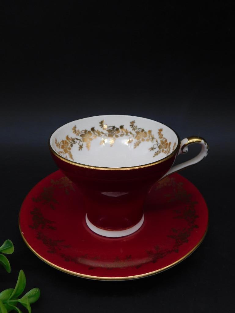 Aynsley England Red Bone China Teacup and Saucer Set: This Aynsley England teacup and saucer set features a deep red design with gold floral embellishments. Made of fine bone china, the set is marked 'Est. Aynsley England Bone China 30' on the underside.
