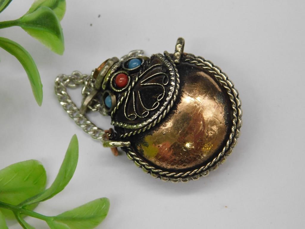 Mixed Metal Snuff Bottle With Gemstone Accents: This mixed metal pendant features a round copper-colored centerpiece encased in an ornate silver-toned frame with braided details. The pendant is adorned with small red, blue, and turquoise stone acce