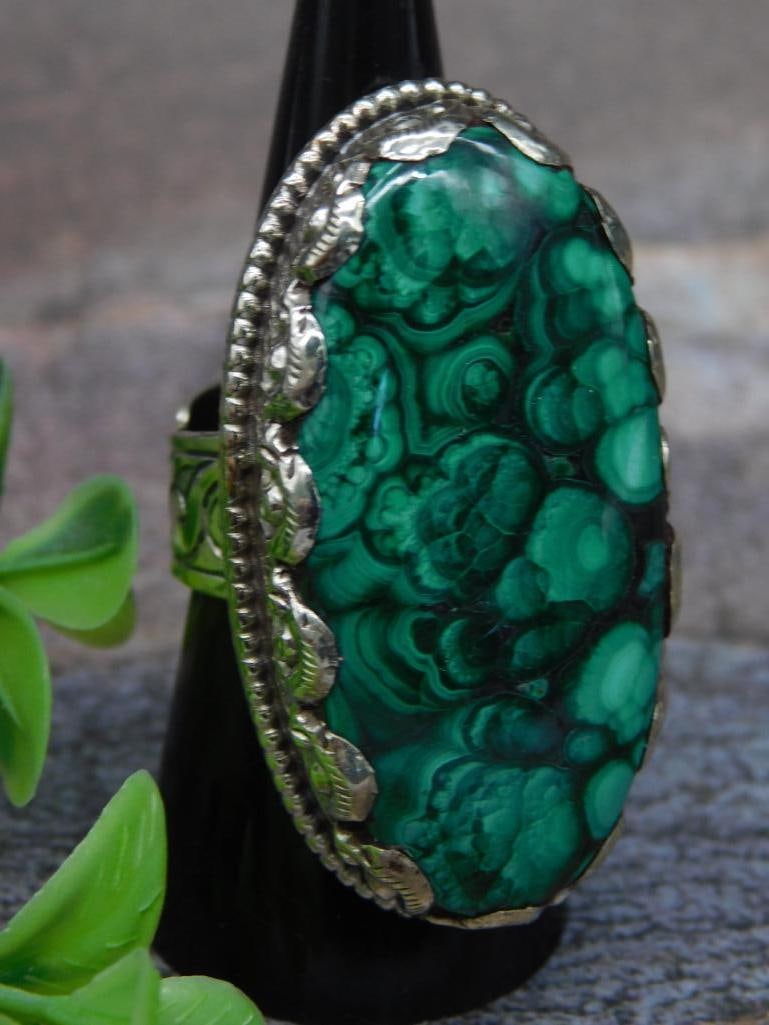 ADJUSTABLE MALACHITE RING WITH INTRICATE TOOLING: Silver-tone ring featuring a large oval malachite-style stone with green swirling patterns. The metal band and underside are intricately engraved. The design showcases detailed craftsmanship, with a s