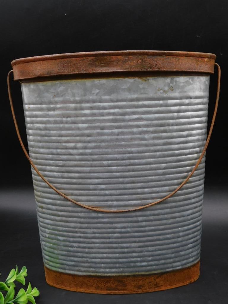 Vintage Galvanized Metal Bucket With Rusted Accents: This is a vintage galvanized metal bucket featuring a ribbed body for added structural strength. It has a rusted metal handle and trim, adding character to its design. The bucket shows signs of wear c