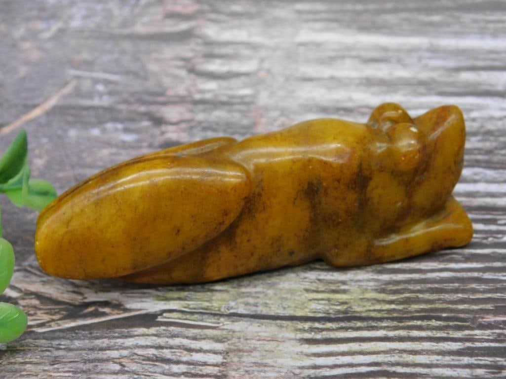 Carved Jade Frog Figurine: Carved Jade figurine depicting a frog with detailed features. The piece has a smooth surface and displays natural amber coloring with marbled variations. Includes a size reference next to a coin for s