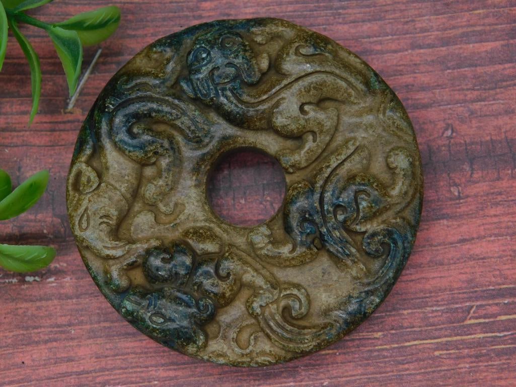 CARVED JADE ROCK STONE LAPIDARY SPECIMEN: - Circular stone disc with a carved design featuring dragon-like motifs. | - Central hole design, typical of traditional bi discs. | - Dark green and brown hues with areas of black shading. | - Materi