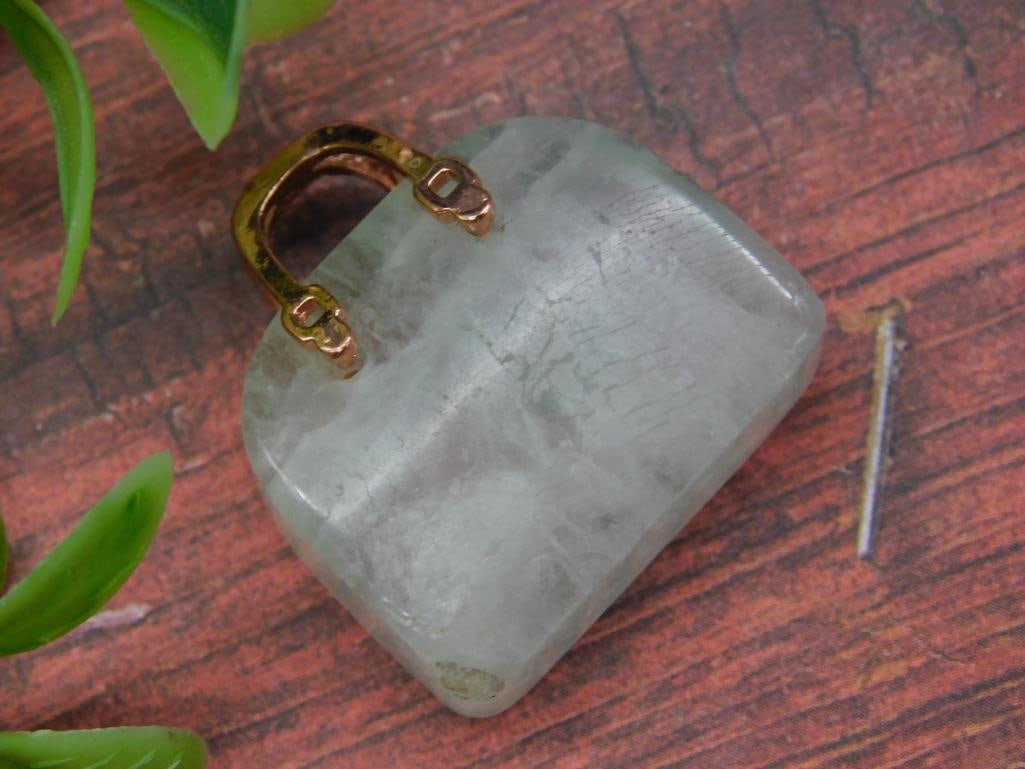 Translucent Stone Pendant with Gold-Tone Bail: This lot features a translucent stone pendant with a subtle green coloration. The pendant includes a gold-tone metal bail shaped like a handle for attachment. Included in the image for scale is a U.S.