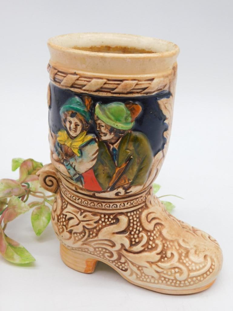 Ceramic Boot-Shaped Vase with Decorative Scenes: Boot-shaped ceramic vase featuring detailed carved patterns and raised, colorful scene designs on both sides. The artwork depicts rustic-style characters in blue, green, and earthy tones. The vase has