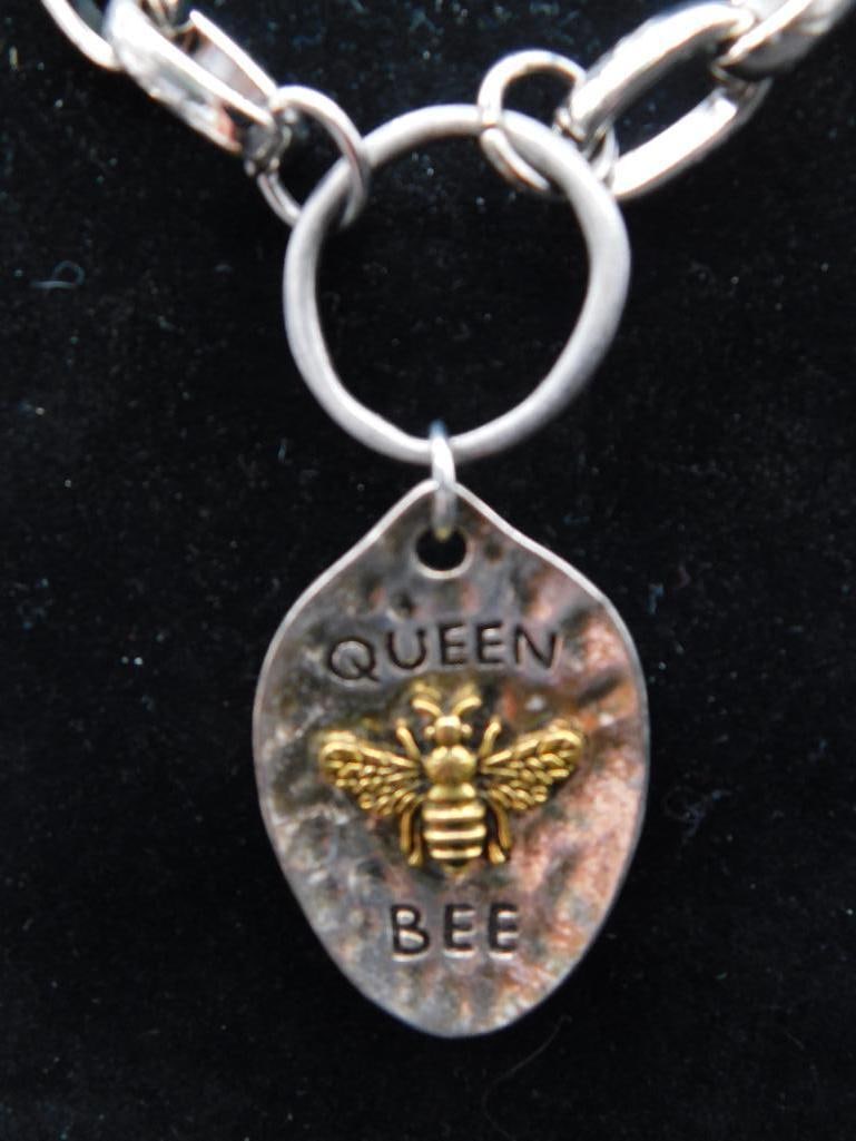 Silver-Tone Chain Bracelet with Queen Bee Pendant: Silver-tone chain bracelet with a distinctive oval pendant featuring a raised golden bee design and the words 'QUEEN BEE'. The bracelet features a sturdy loop clasp for secure wear. Presented on a dis