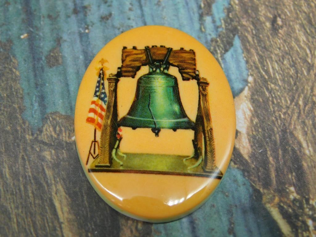 Oval Cabochon Featuring Liberty Bell and U.S. Flag Design: - Oval-shaped pin featuring an image of the Liberty Bell with an adjacent U.S. flag. | - The background appears to have a muted yellow tone, highlighting the designs. | - Pinback likely made of metal 