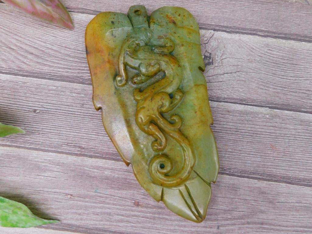 Carved Green Jade Leaf Pendant With Embossed Design: Carved green stone pendant in the shape of a leaf with spiral and embossed design. Features tones of green and brown. Includes a small hole for stringing or attaching to jewelry. The pendant weighs 80