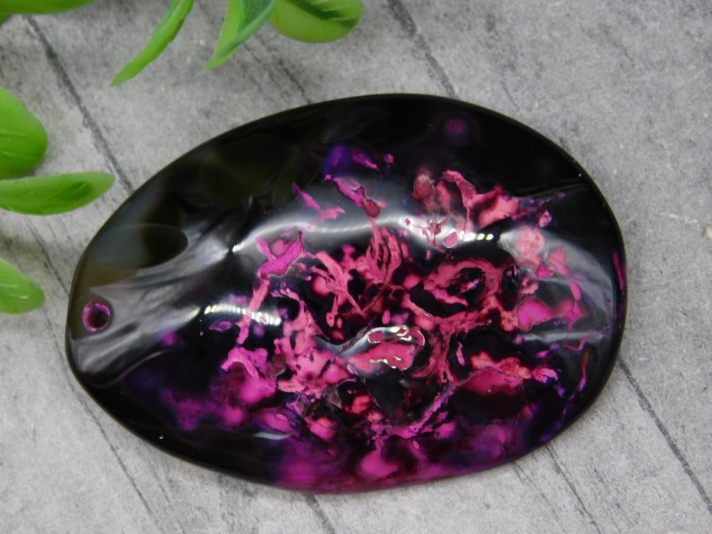Oval Pendant With Pink and Purple Pattern: Oval-shaped pendant featuring a black background with vibrant pink and purple patterns. Includes a small pre-drilled hole ideal for stringing or attaching. Estimated weight is 27 grams. Measures appro