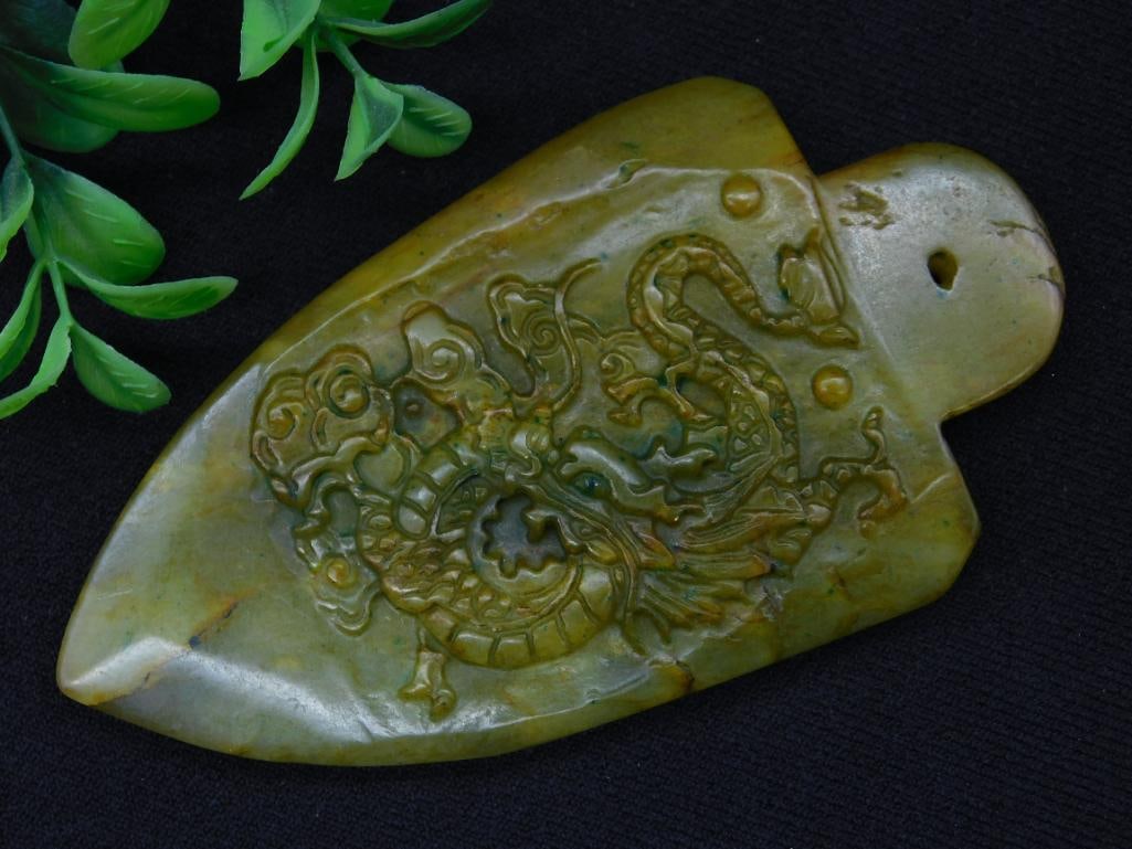 Carved Green Jade Pendant Featuring Dragon Motif: Carved green stone pendant with an intricate dragon motif. The item measures approximately 80 grams and has a hole at the top for hanging or attaching. It shows detailed craftsmanship with a beautiful
