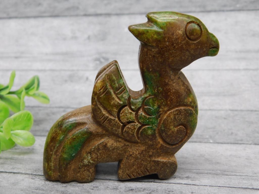 Carved Jade Figurine of a Mythical Creature: Carved stone figurine depicting a mythical creature, possibly resembling a dragon or gryphon. Features detailed carving with intricate wing and tail designs. Surface showcases natural green and brown