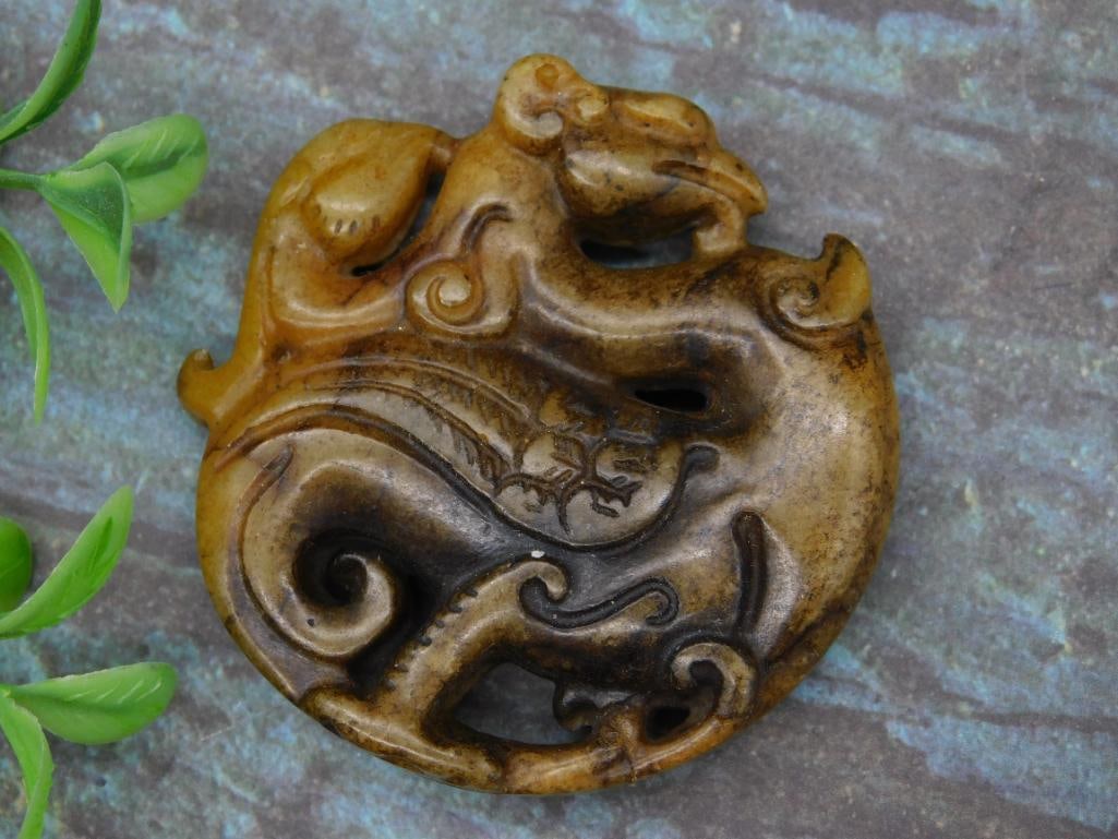 Carved Jade Stone Dragon Pendant: A carved brown stone pendant featuring a dragon design. It appears to have detailed ornamental patterns and a circular form. The item's scale is shown with a U.S. quarter for size reference, though sp