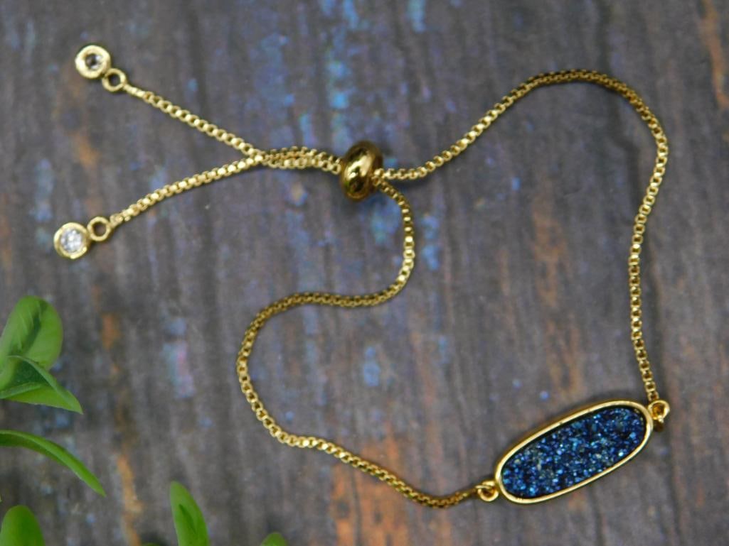 Gold-Plated Druzy Bracelet with Adjustable Chain: Gold-plated bracelet featuring an adjustable chain for a custom fit. Centerpiece includes an oval druzy stone with a sparkling blue finish. Design incorporates a sliding bead clasp and CZ accents on t