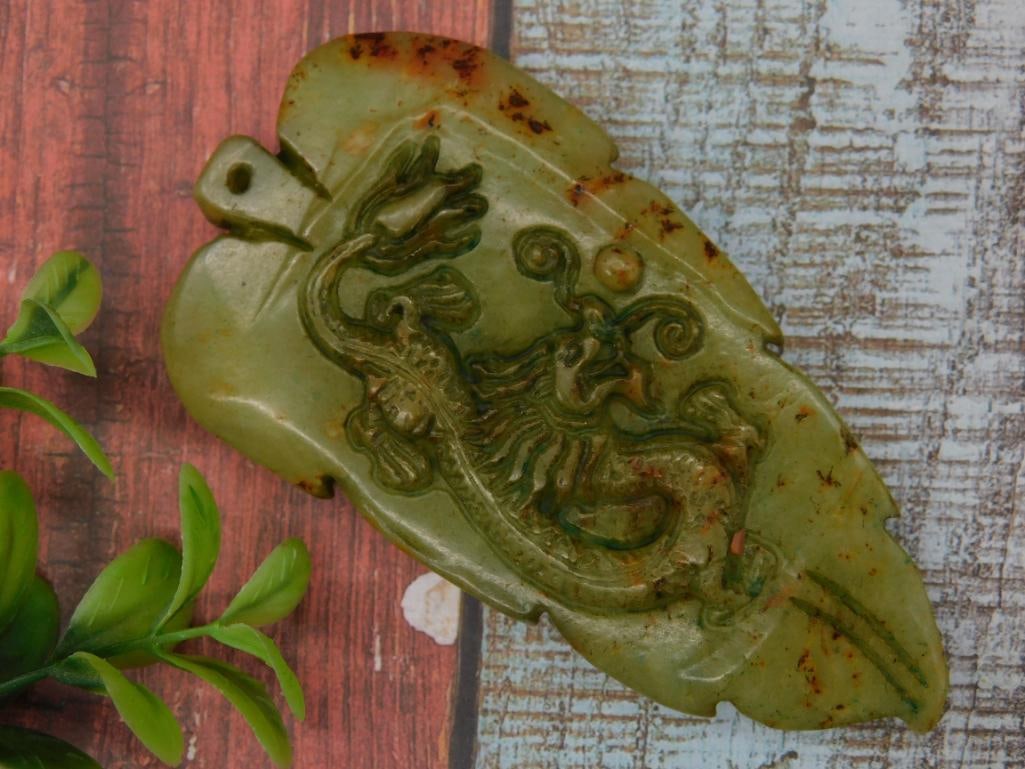 Carved Green Jade Pendant With Dragon Motif: Green stone pendant featuring a carved dragon motif, thoughtfully textured and detailed. Includes a small hole for hanging or stringing as a pendant. Dimensions visible in the image indicate a length