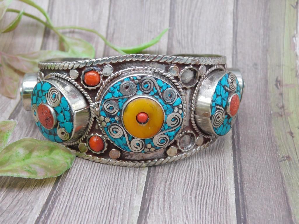 Decorative Metal and Stone Cuff Bracelet with Turquoise and Red Coral: A metal cuff bracelet featuring detailed decorative elements and inlaid stones. This design includes turquoise, red, and yellow stone accents with intricate silver embellishments. It measures approxim