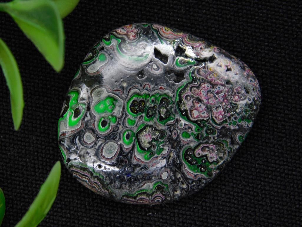 FORDITE MOTOR CITY AGATE CABOCHON ROCK STONE LAPIDARY SPECIMEN: A single polished mineral specimen showcasing vivid green, black, and other swirled patterns. The rock features dynamic natural formations and varied shades, making it a visually appealing piece. Coin