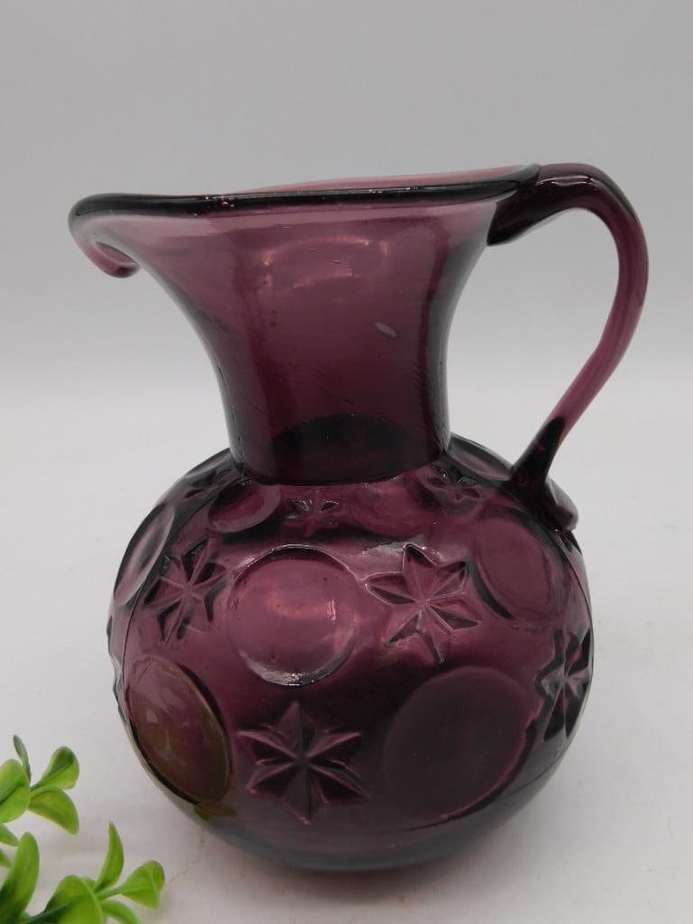 AMETHYST PITCHER VINTAGE ANTIQUE: Small purple glass jug featuring a star and circle pattern design. It has a single curved handle and a flared spout. The item shows expected wear on its underside, typical for glassware. Its approxima