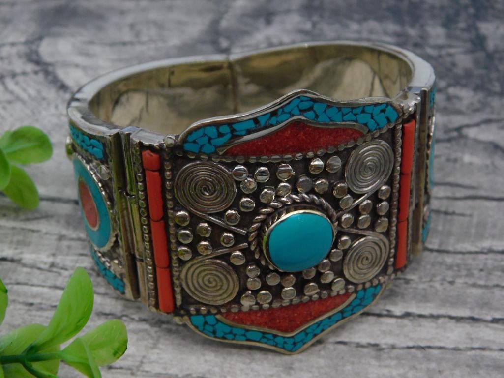 Turquoise and Red Coral Inlay Silver Bracelet: - Silver bracelet featuring turquoise and coral inlay designs. | - Intricate raised details including spiral and beaded motifs. | - Hinged construction for easy wear. | - Vibrant blue, red, and silver