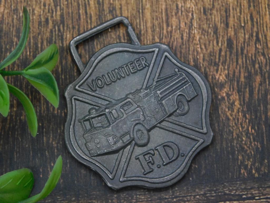 Volunteer Fire Department Watch Fob: Metal watch fob featuring a fire department design. | - Includes text 'Volunteer' and 'F.D.' with a fire engine design in the center. | - Shaped in a maltese cross style, common with firefighting imag